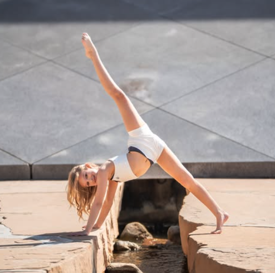 A young dancer wearing white performing a handstand on a rock with one leg raised, showcasing strength and flexibility against a modern architectural background.